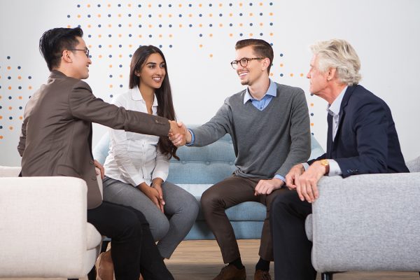Smiling young business men shaking hands. They and their partners are sitting in armchairs and sofa in lounge.